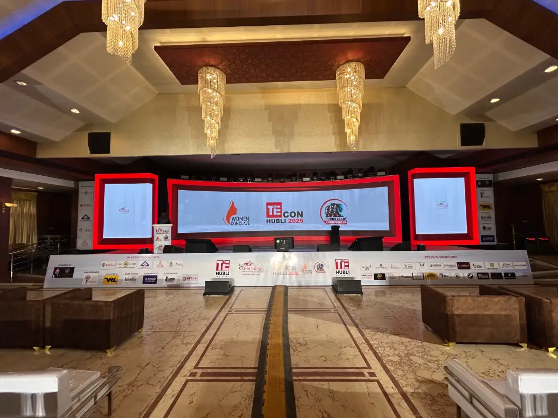 A red-and-white TiE conference stage with wide LED screens under chandeliers
