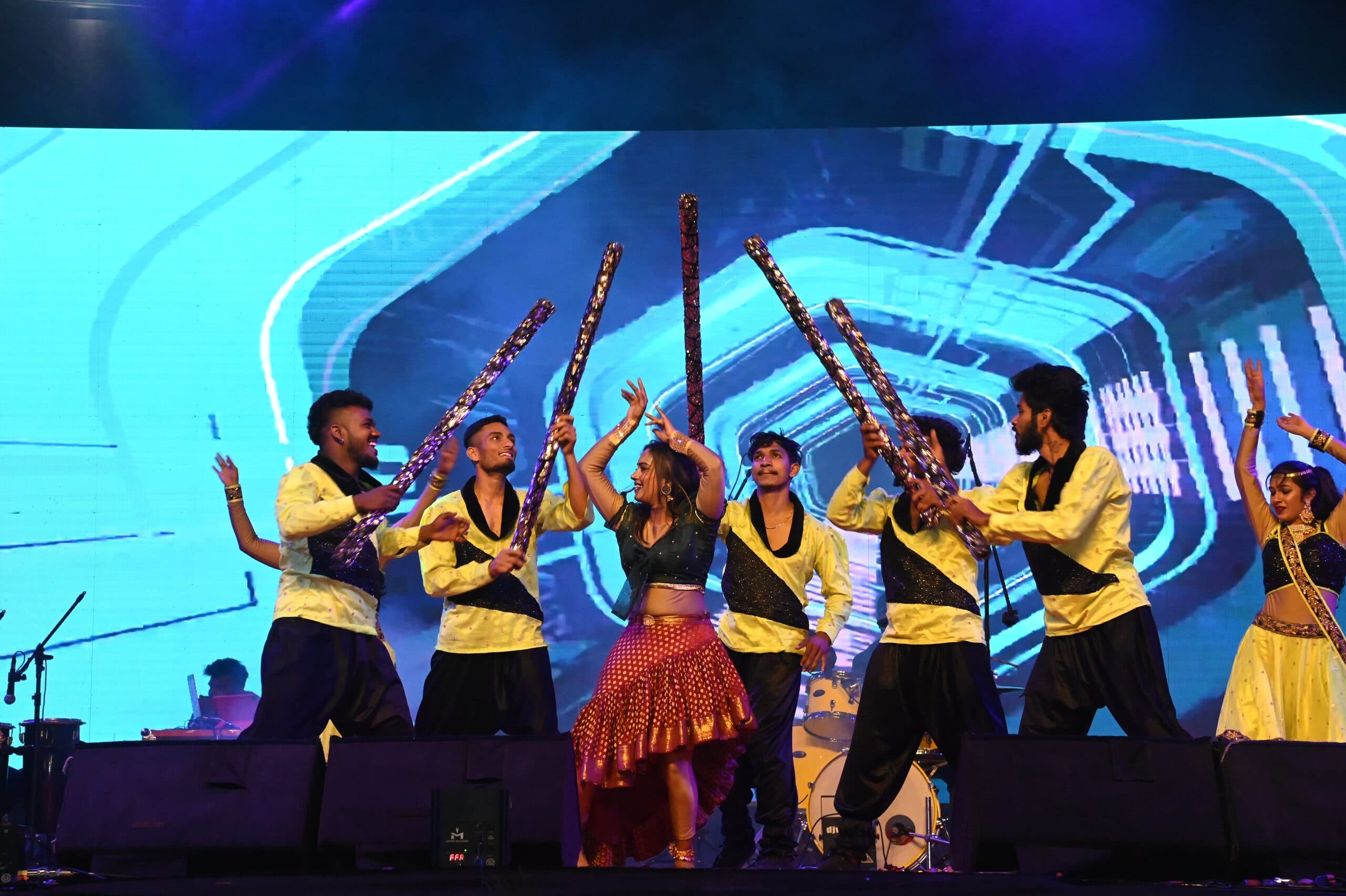 A folk-fusion troupe raising kolata sticks in front of a curved cyan LED backdrop