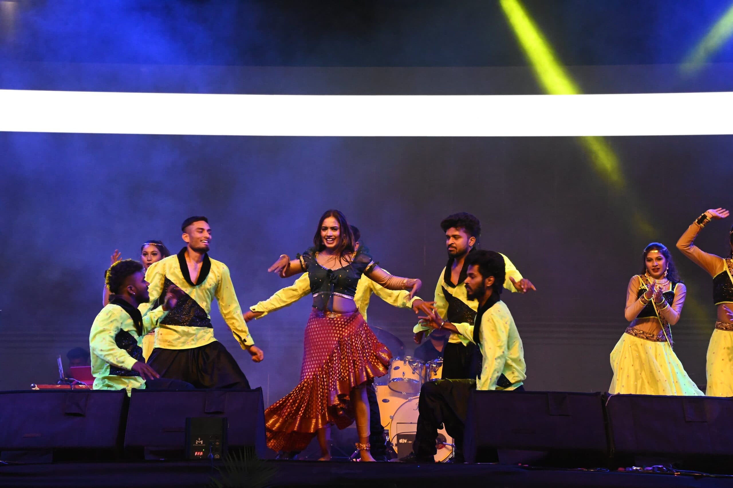A folk troupe in yellow and red mid-routine on the Ugadi festival stage under blue and gold beams