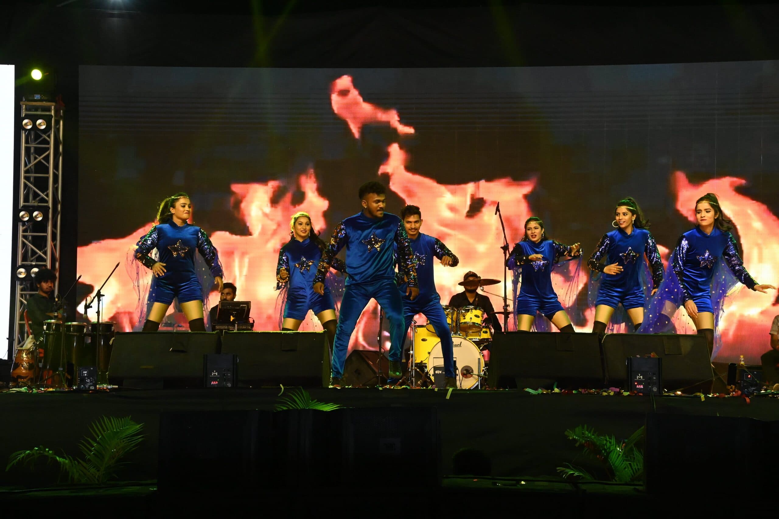Dancers in blue across a wide stage against a flame-graphic LED wall, heads of the audience in silhouette