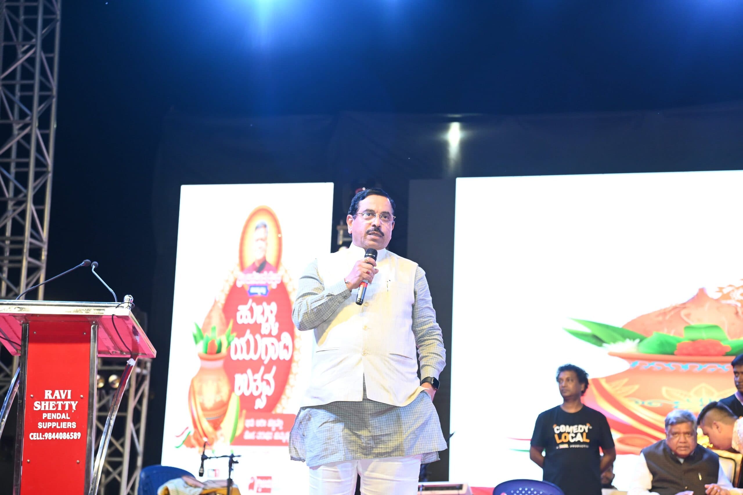 Speaker addressing the crowd from the lectern beside the Hubballi Yugadi Utsava banner