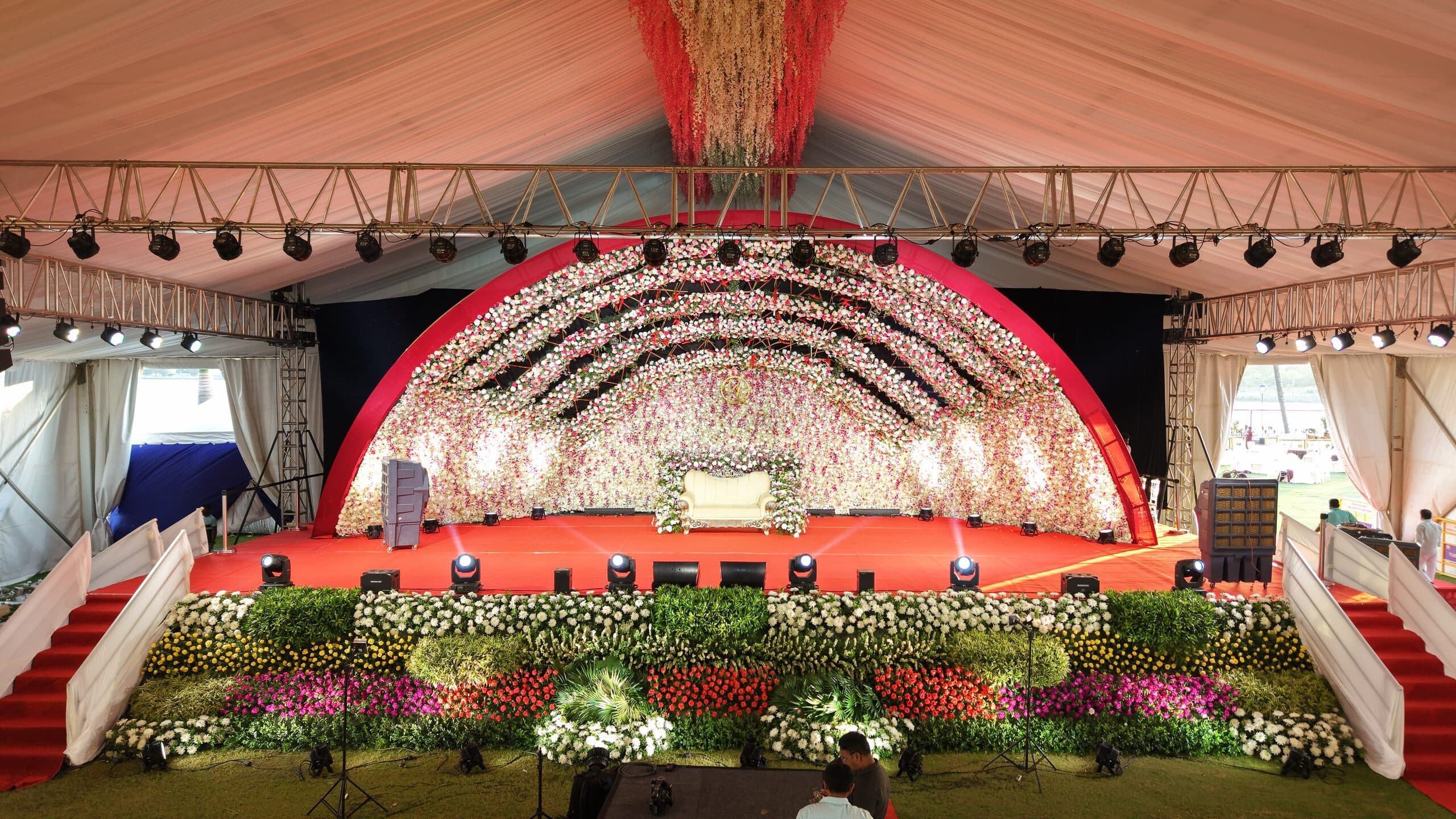 An arched floral reception stage at the head of a red-carpet aisle inside a marquee