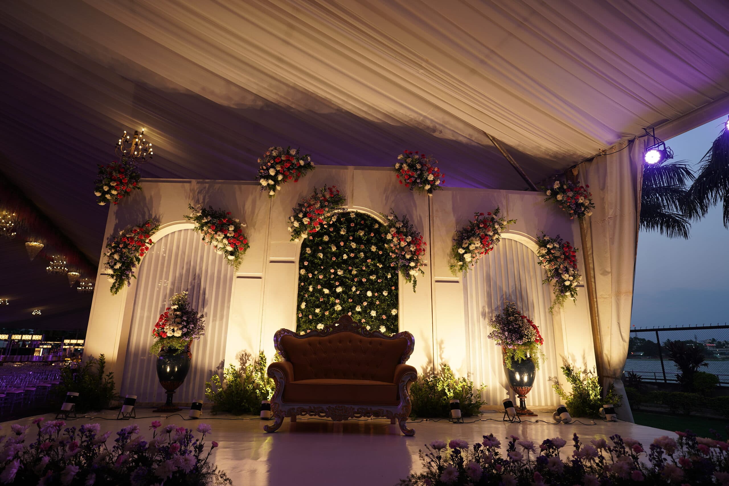 An evening reception stage in ivory drape and flowers, uplit purple, with a single carved chair
