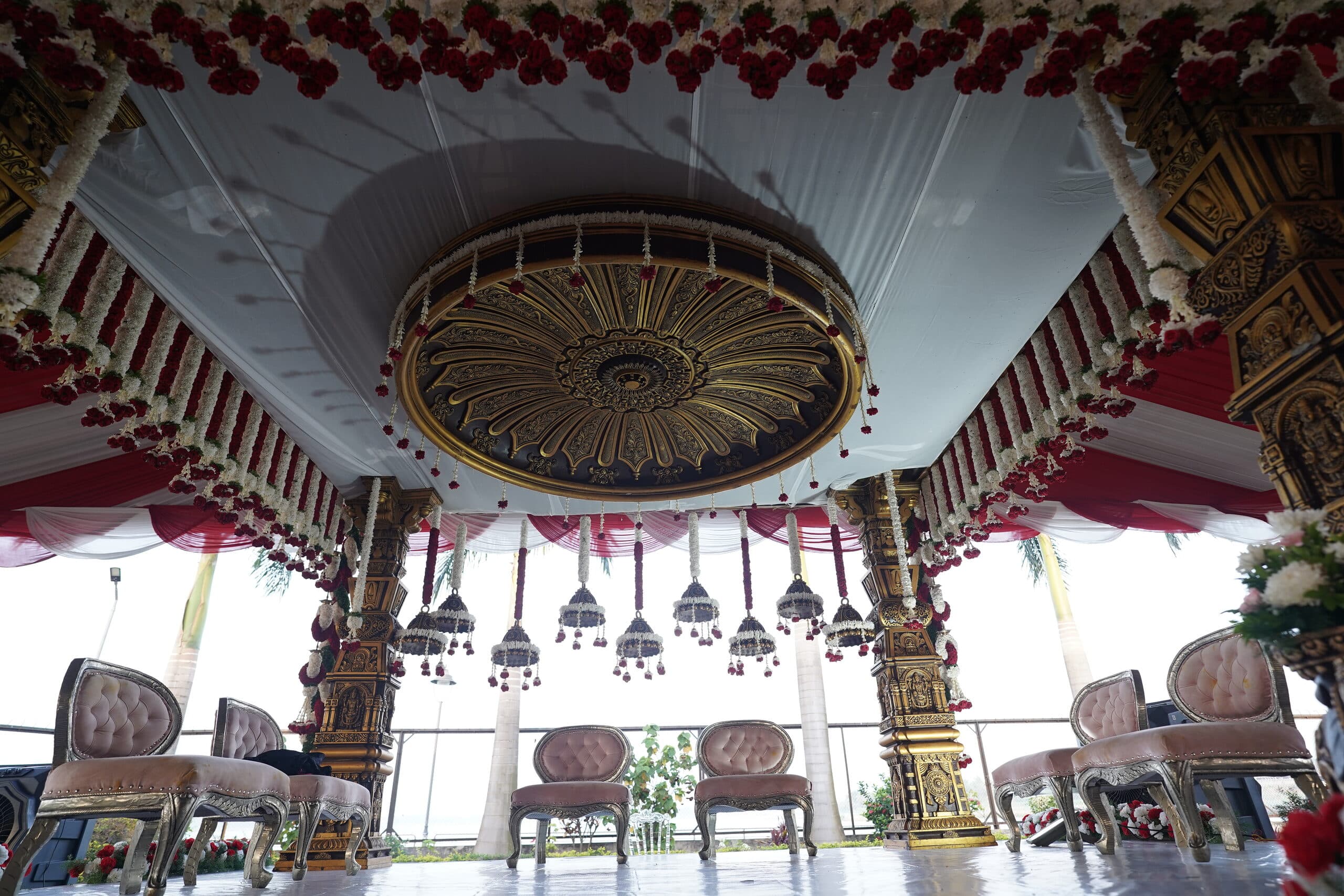A carved mandap interior hung with rose garlands beneath a gilded dome ceiling