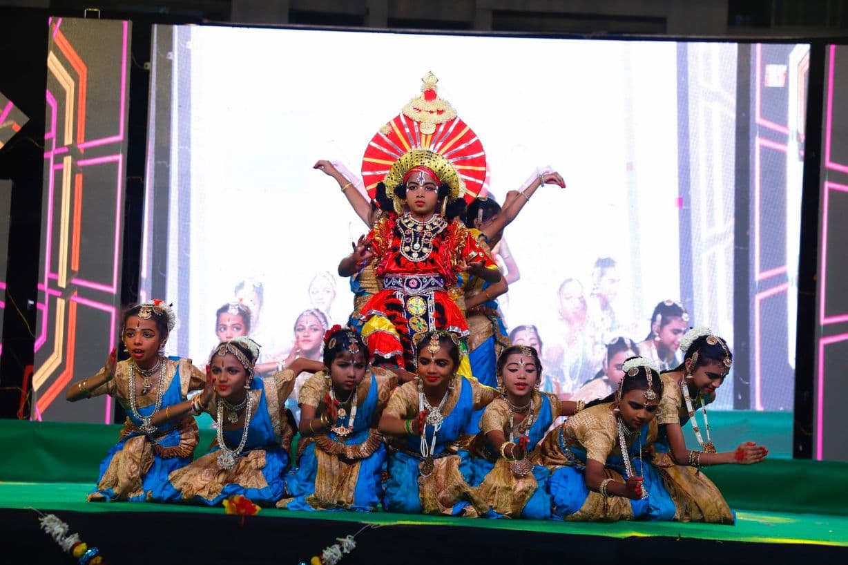 Costumed children seated around a crowned deity tableau on a green-lit college stage