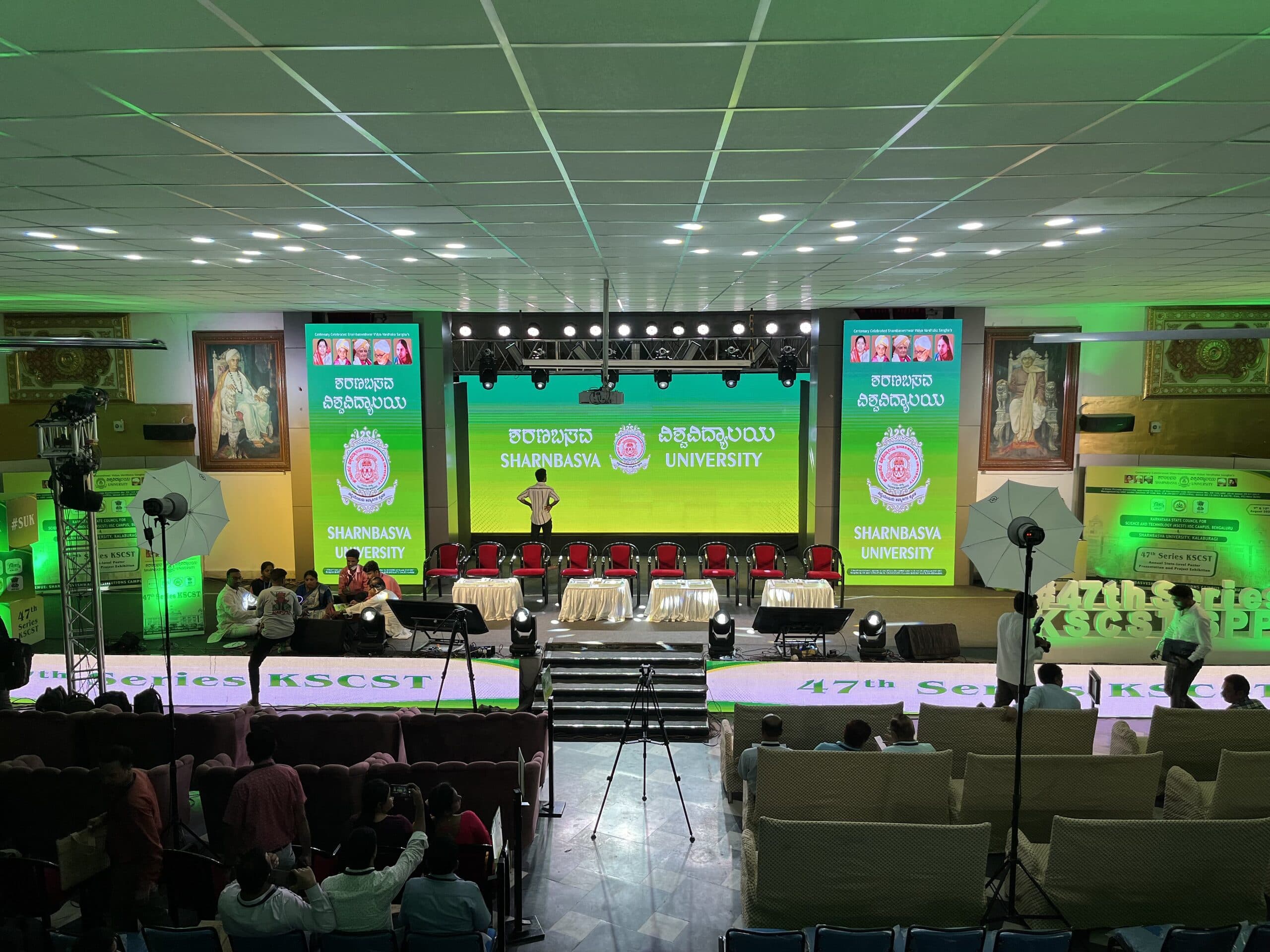 Green-lit university stage under a full-width LED wall, flanked by portrait banners
